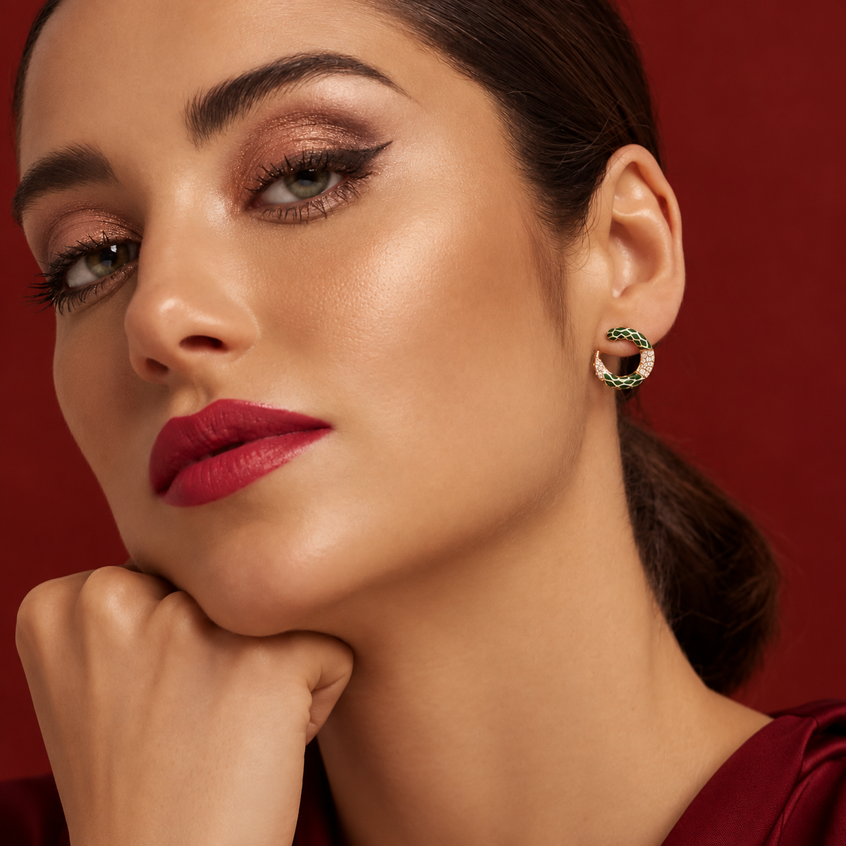 A close-up portrait of a woman wearing a textured green and gold hoop earring, set against a solid deep red background.
