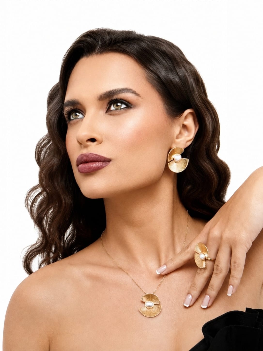 A woman with dark wavy hair models a matching gold jewelry set featuring a disc-shaped pendant necklace, a matching drop earring, and a ring with a pearl accent, set against a plain white background.