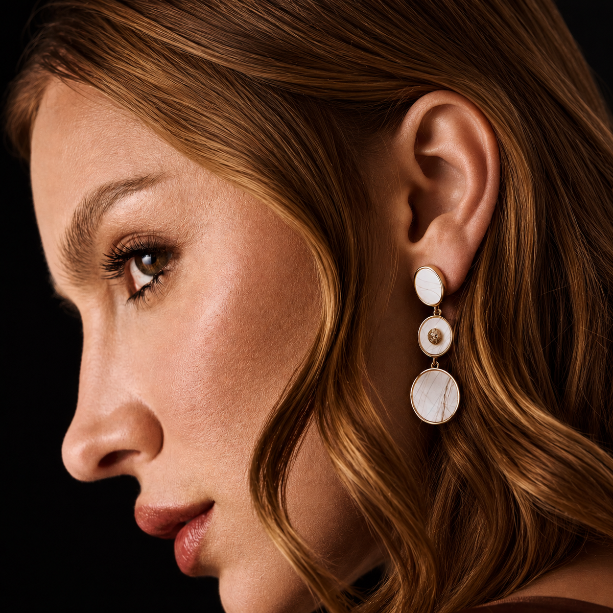 A close-up side profile of a woman with wavy light brown hair wearing a gold and white mother-of-pearl drop earring 